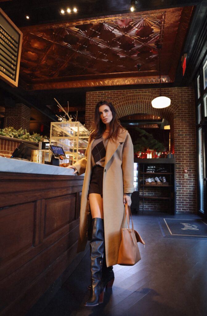 Misha Skova standing in a rustic café wearing a camel wool coat, argyle knit sweater, black shorts, and over-the-knee leather boots with a tan leather tote bag.