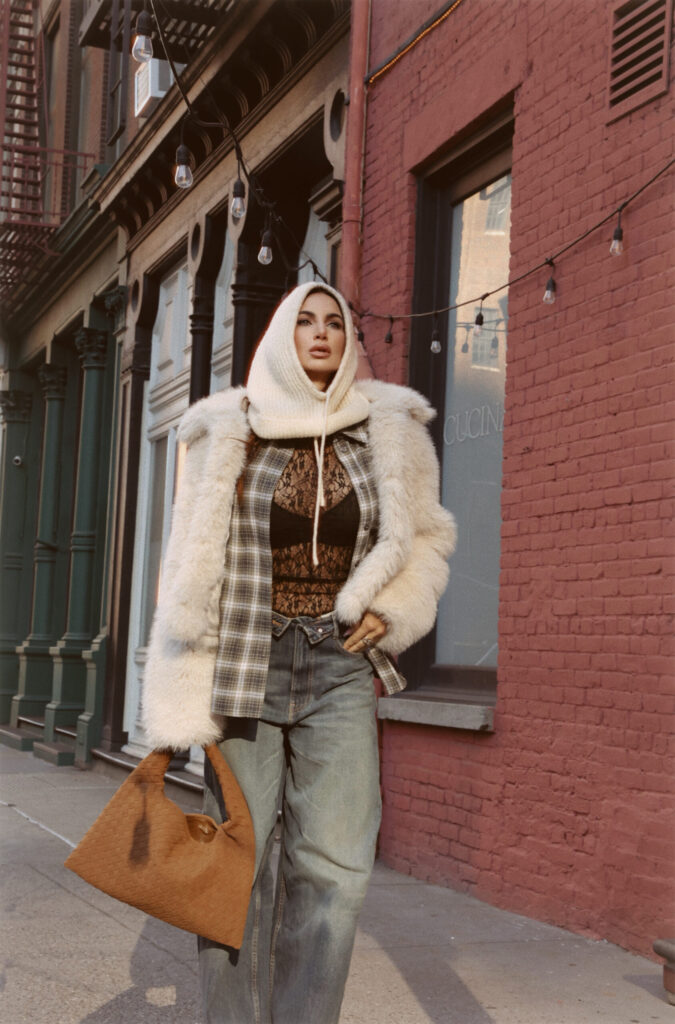 Misha Skova modeling winter fashion trends 2025-2026 in an urban setting, wearing a cream faux fur coat layered over a plaid shirt and black lace top with wide-leg denim jeans.