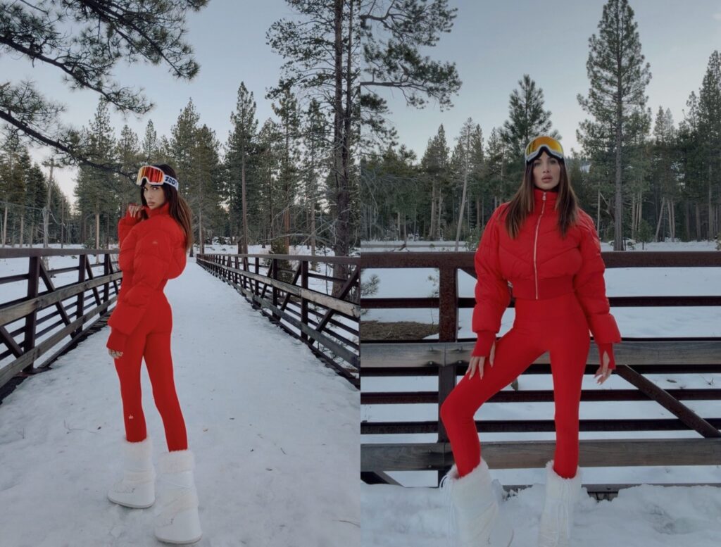 Misha Skova in a bright red winter outfit by Alo, including a puffer jacket, fitted pants, white fur boots by Moon Boot, and ski goggles by 100%, stands on a snowy wooden bridge surrounded by pine trees.
