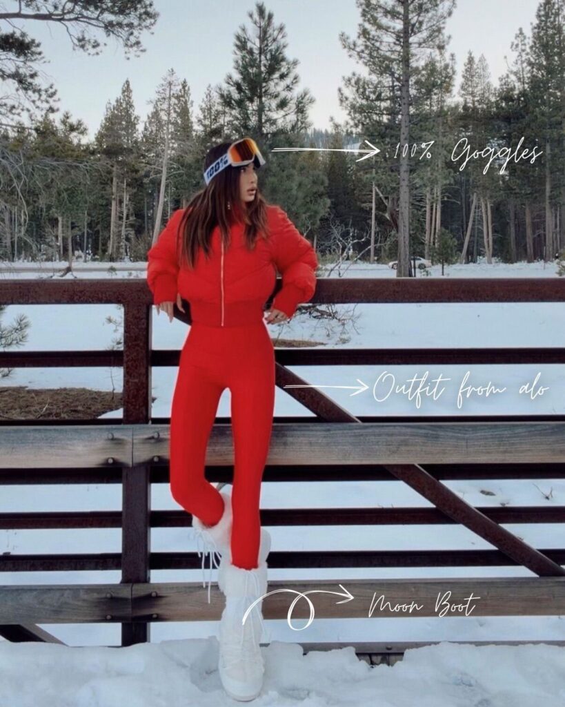 Misha Skova in a bright red outfit by Alo and white snow Moon boots stands on snow in front of a wooden fence with pine trees behind her. She wears ski goggles by 100% and poses with her hands on her hips. Text labels highlight her goggles, outfit, and boots.