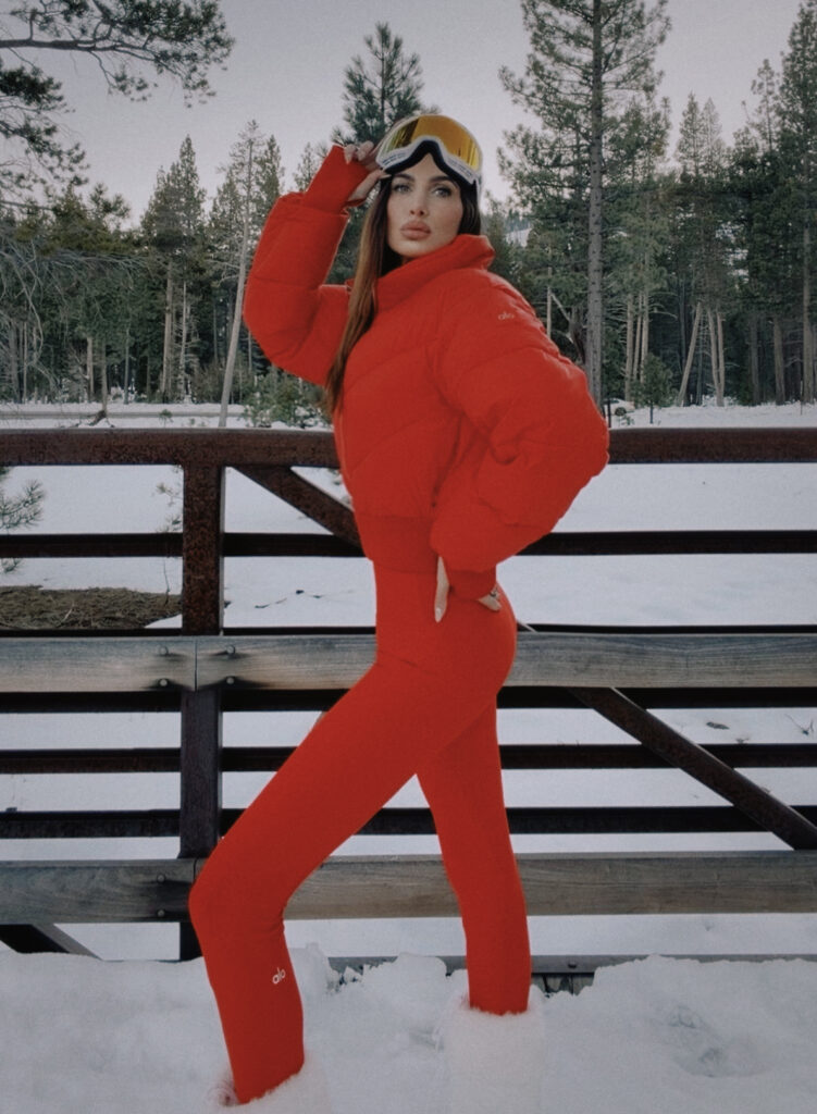 Misha Skova in a bright red winter outfit by Alo, including a puffer jacket, fitted pants, white fur boots by Moon Boot, and ski goggles by 100%, stands on a snowy wooden bridge surrounded by pine trees.