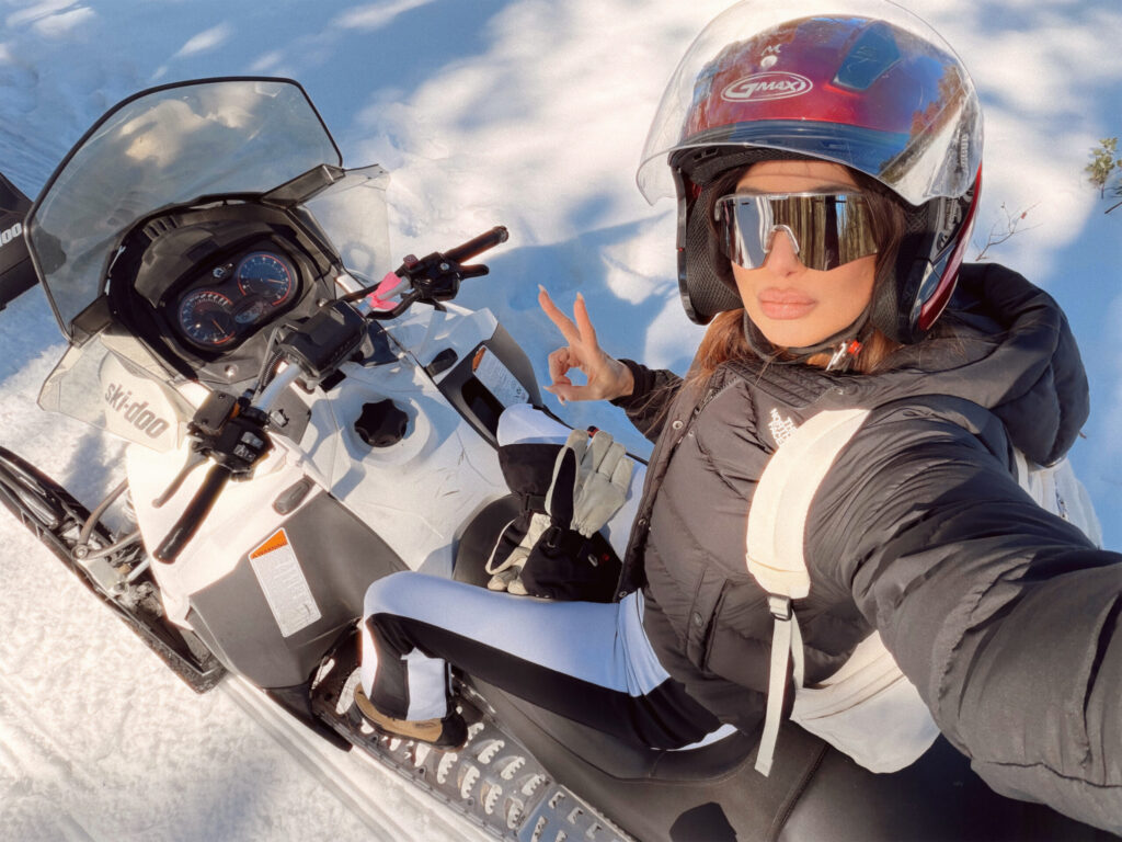 Misha Skova wearing a helmet, sunglasses, and winter clothing sits on a snowmobile in the snow, taking a selfie and making a peace hand gesture.