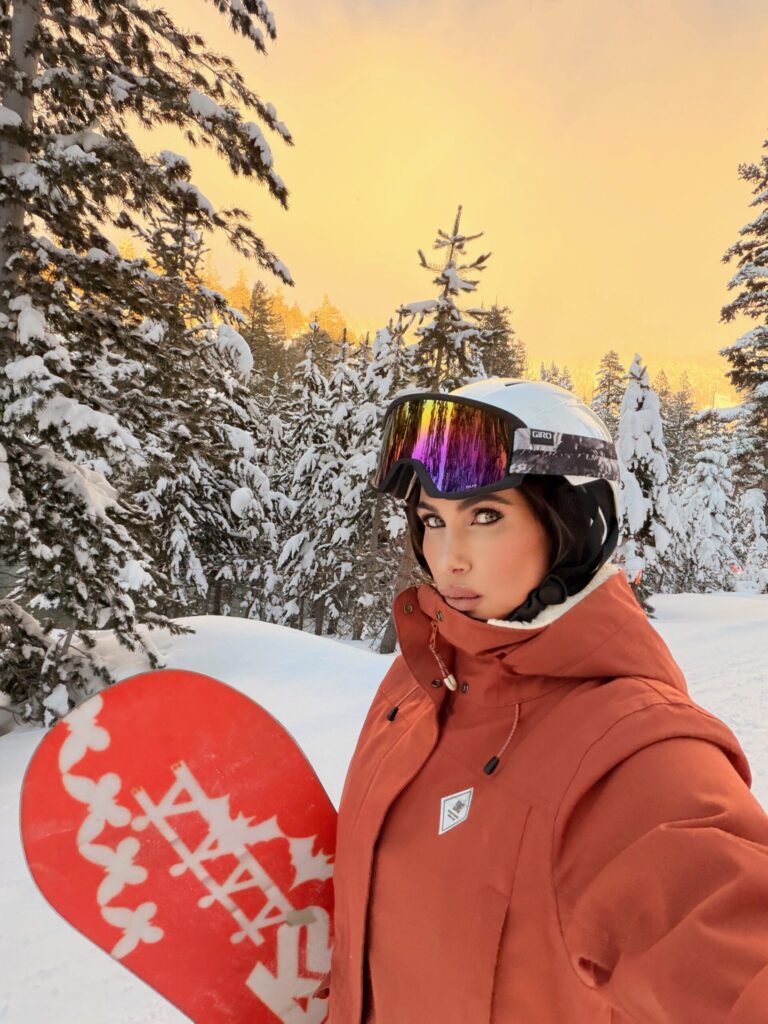Misha Skova in a red jacket and helmet holds a red snowboard, standing on a snowy slope surrounded by snow-covered trees at sunset, with a vibrant orange sky Lake Tahoe in the background.
