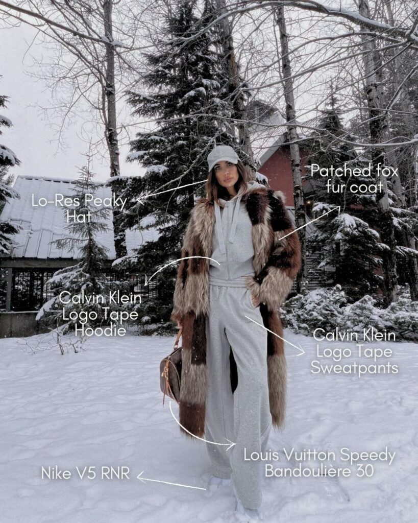 Misha stands in a snowy landscape wearing a gray Lo-Res Peaky hat, a patched fox fur coat, and a gray Calvin Klein logo tape hoodie and sweatpants. They hold a Louis Vuitton Speedy Bandoulière 30 bag and wear white Nike V5 RNR shoes. Snow-covered trees and a house are in the background.