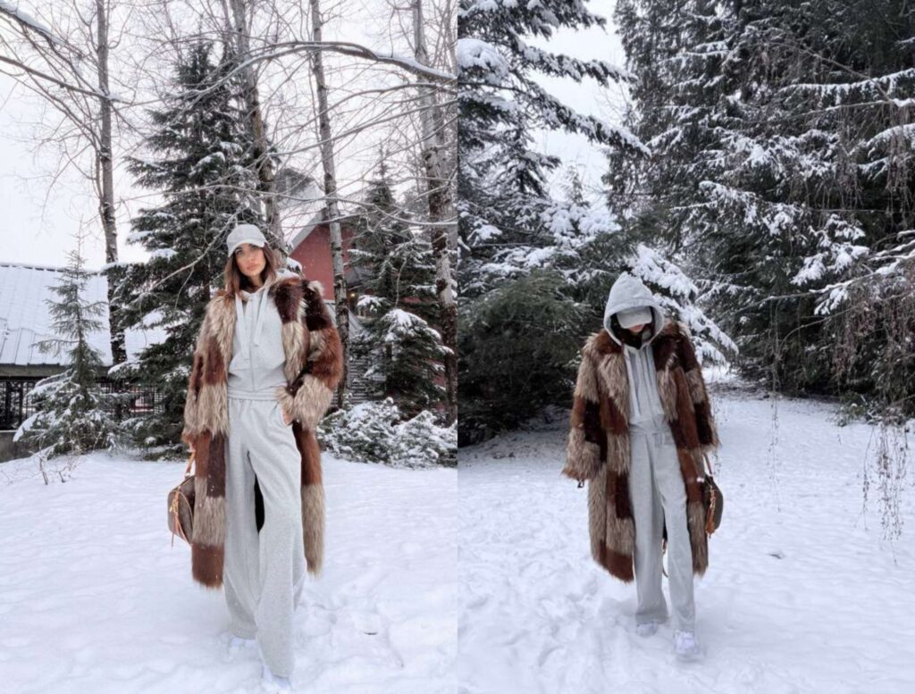 Misha Skova wearing a long fur coat, light gray hoodie, sweatpants, and white sneakers poses in a snowy outdoor setting surrounded by trees and a red building in the background.