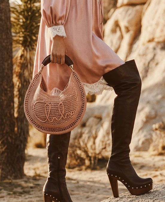 A person in a pink dress with lace cuffs and black knee-high platform boots stands outdoors, holding a woven round handbag with Chloé written on it. Desert plants and rocks are in the background. Chloé Outfit Inspiration