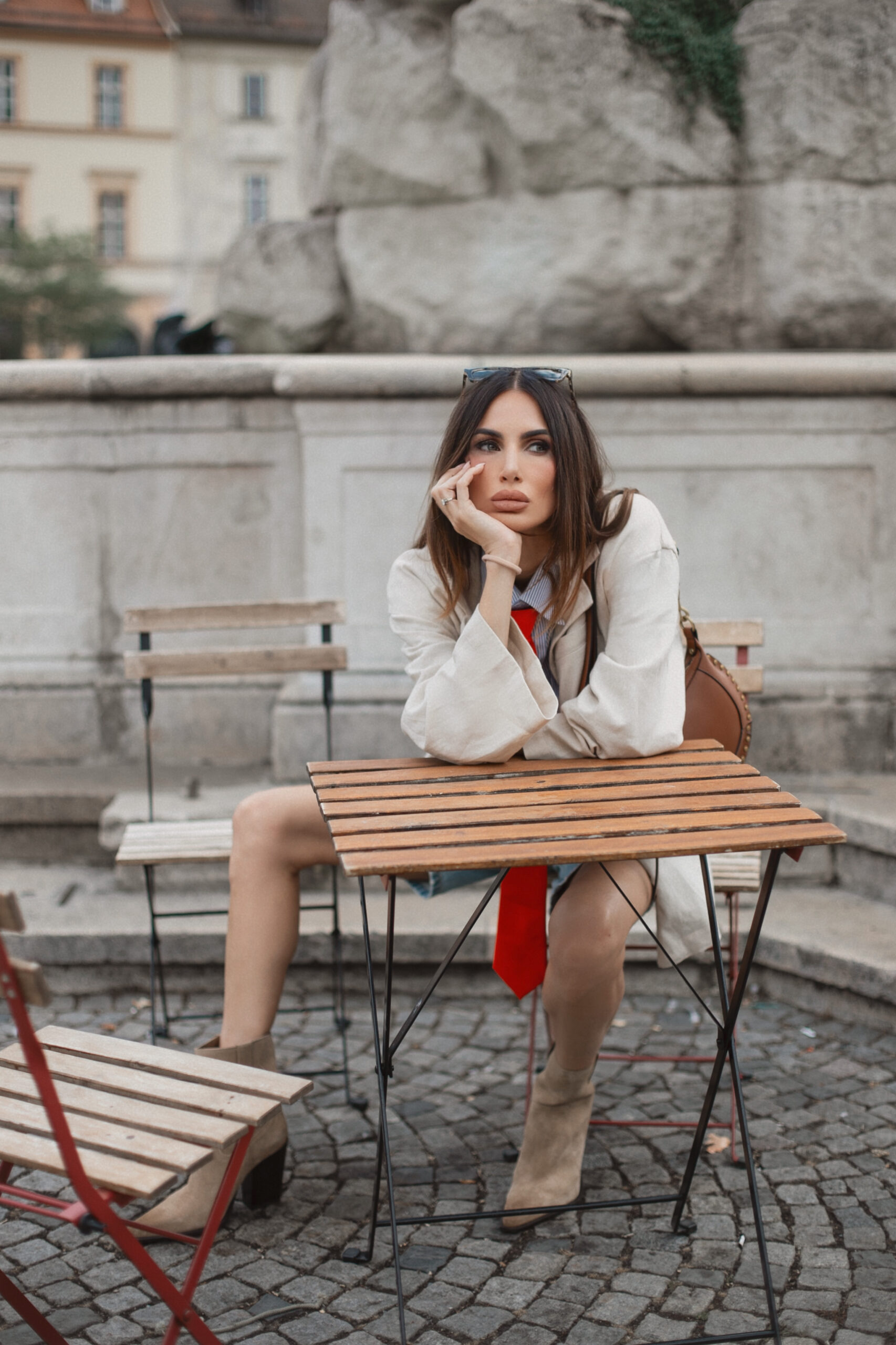 Woman seated at an outdoor bistro table in Brno wearing sunglasses on her head, a striped shirt with orange tie, beige blazer, and denim shorts