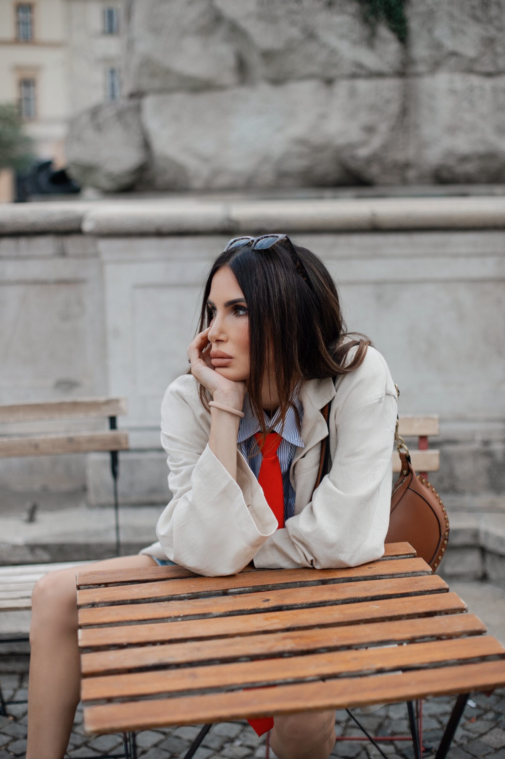 Woman resting her chin on her hand at an outdoor bistro table, wearing a striped shirt, orange tie, and beige blazer