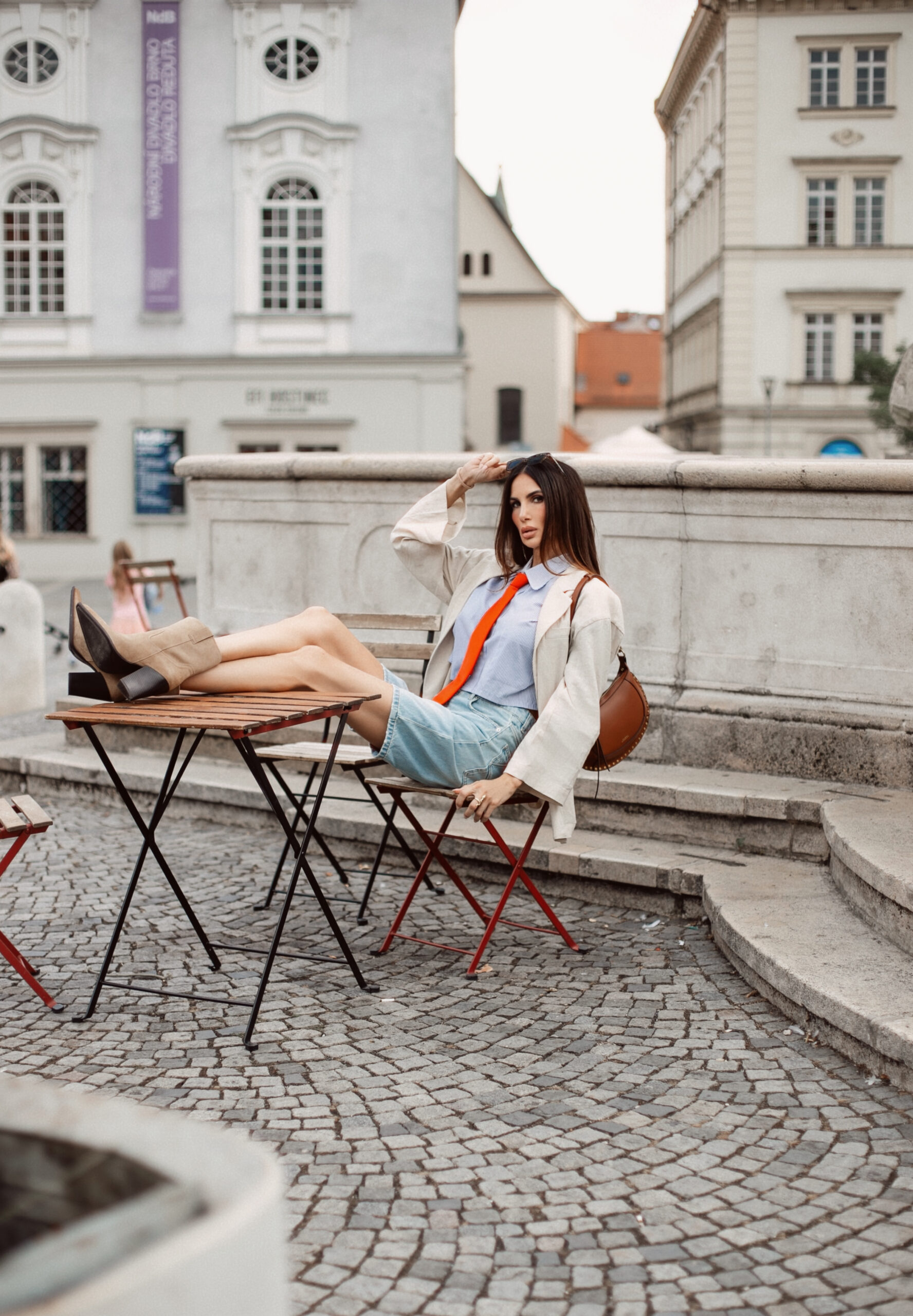 Woman with feet propped up on a bistro table, wearing an oversized beige blazer, orange tie, striped shirt, and denim shorts in a Brno square