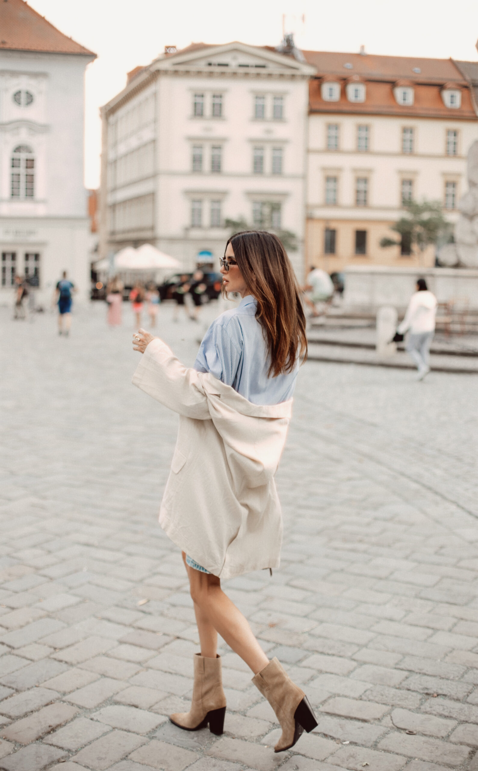 Back view of a woman shrugging off her oversized beige linen blazer over a blue striped shirt in a European cobblestone square