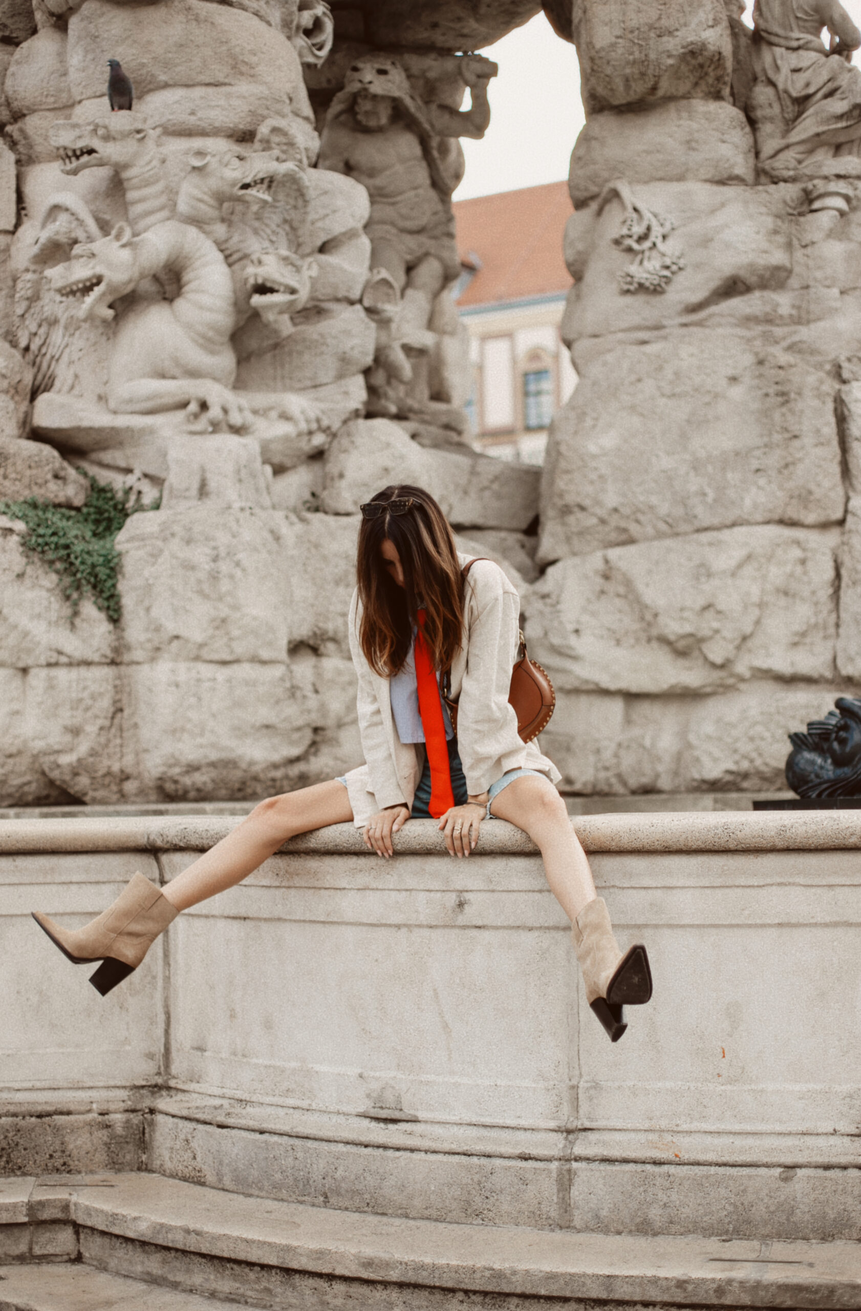 Woman sitting on a stone fountain ledge with legs dangling, wearing beige linen blazer and tan suede boots, ornate gargoyle statues behind her