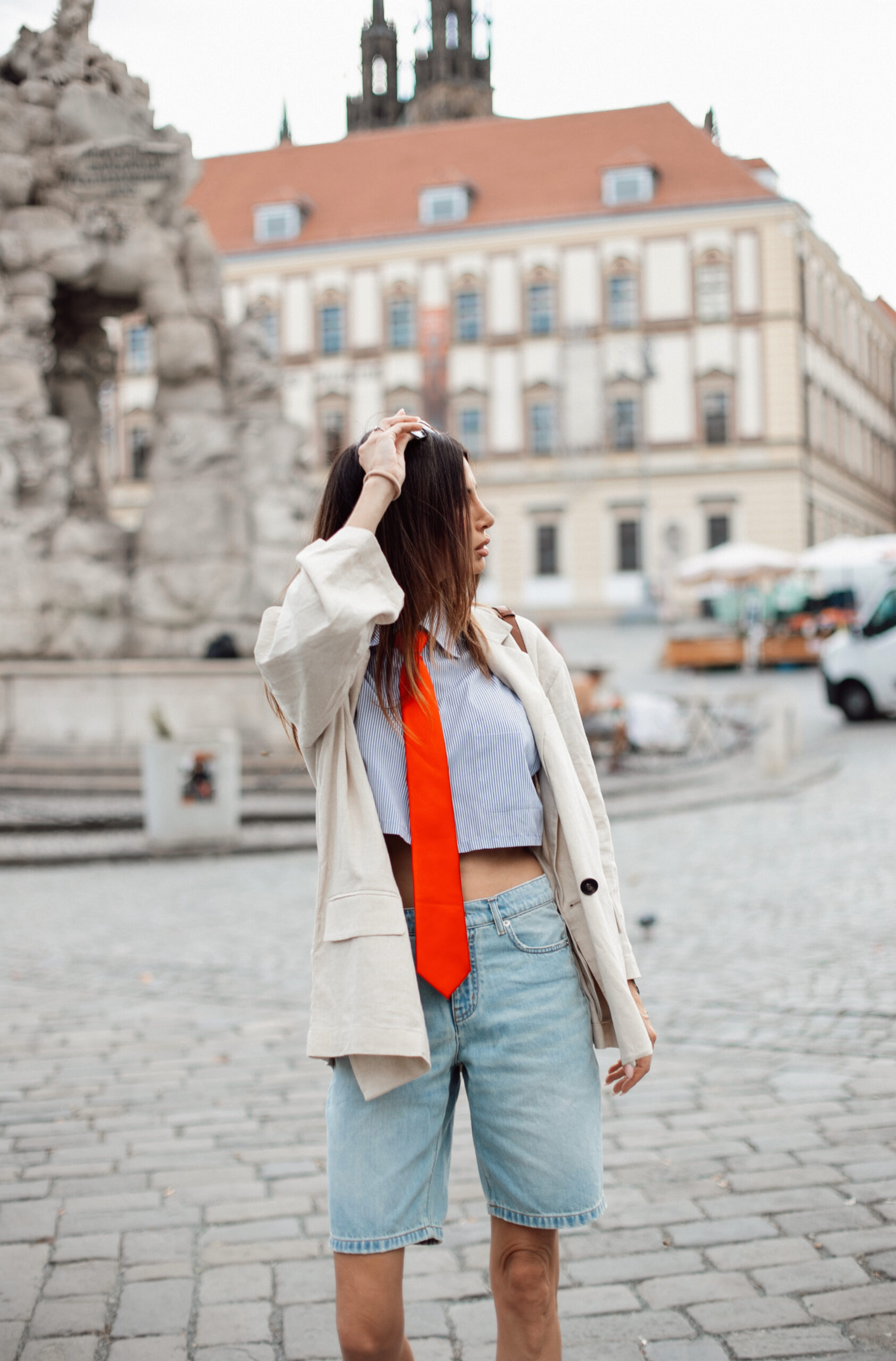 Side profile of a woman adjusting her hair, wearing a striped shirt with an red silk tie under an oversized cream blazer and denim shorts.